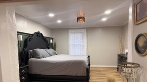 A bedroom with a bed, a nightstand, a dresser, a chandelier, and a window with sheer curtains.