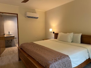 Ein Hotelzimmer mit Bett, Nachttisch, Lampe, Klimaanlage und einem Badezimmer mit Waschbecken und Spiegel.