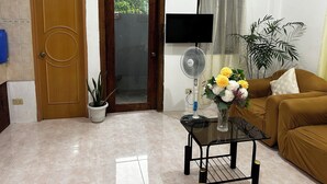 Una sala de estar con un sofá, una mesa con un ventilador, un televisor, una puerta y una planta en maceta.
