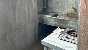 Un espacio reducido y cerrado con un muro de concreto, una planta en maceta, una pequeña mesa con estufa a gas y un estante de metal.