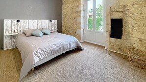 A bedroom with a stone wall, a bed with a white headboard, a large window, and a flat-screen TV mounted on the wall. - Charming estate with swimming pool - Group gîte for 15 people. (Castéra-Verduzan)