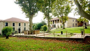 A stone building with a tiled roof, surrounded by a grassy area with trees and a wooden deck. - Charming estate with swimming pool - Group gîte for 15 people. (Castéra-Verduzan)