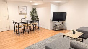 Un salon avec un plancher en bois, une table à manger avec des chaises, un canapé gris, un téléviseur posé sur un meuble et une plante.
