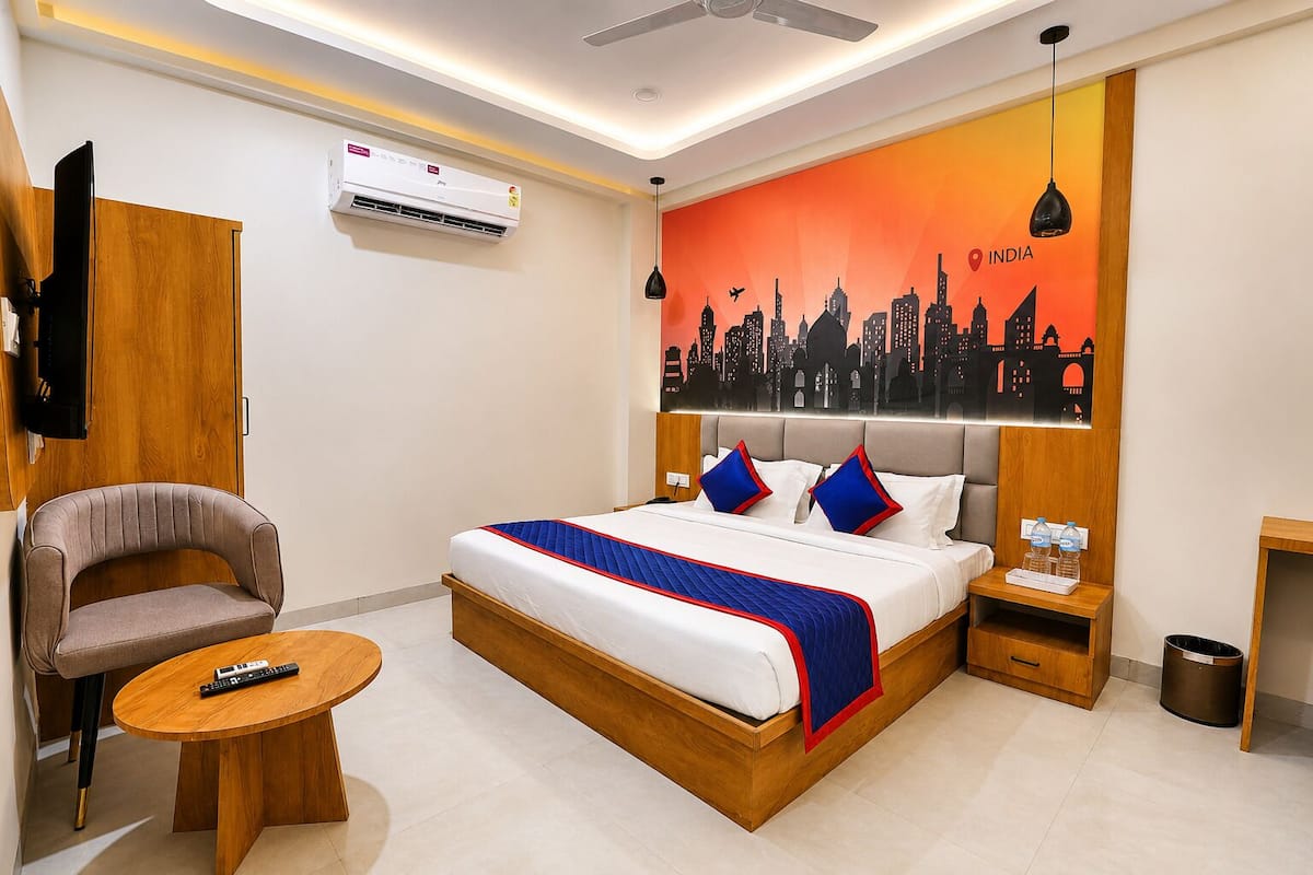 A hotel room with a bed, a chair, a small table, an air conditioning unit, a wall-mounted TV, a ceiling fan, and a cityscape mural.