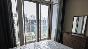 A bedroom with a large window, a bed with a plastic cover, a dresser, and a mirror.
