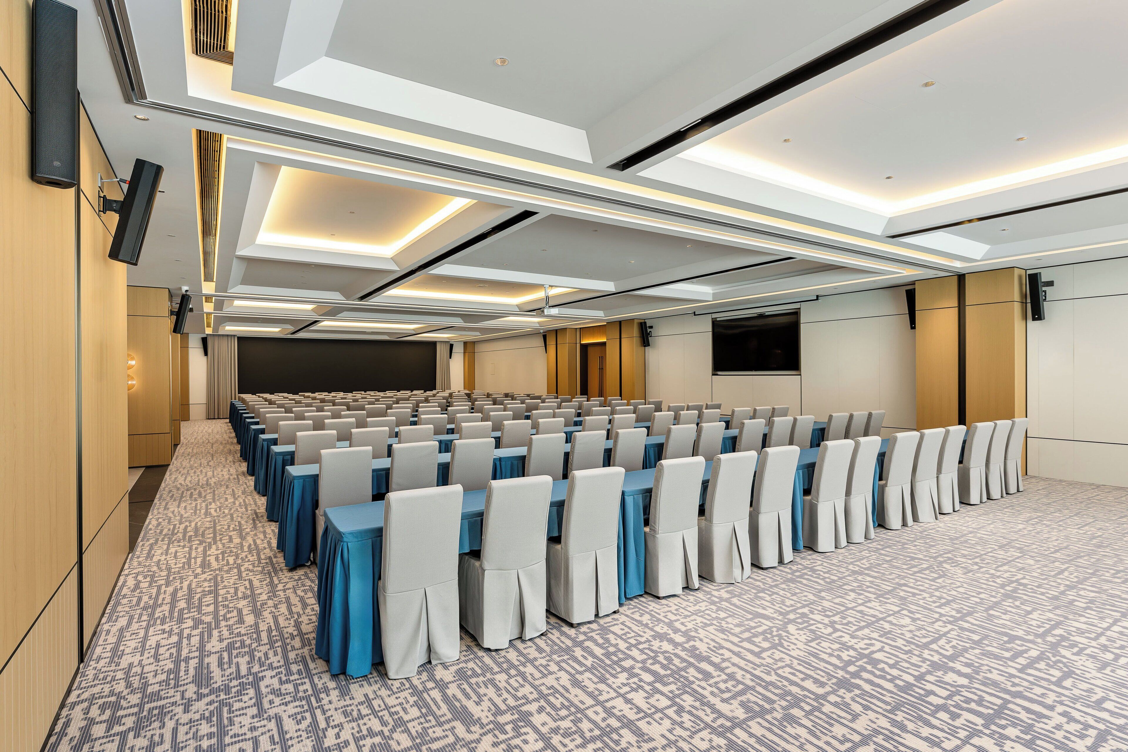 A conference room with rows of tables and chairs, a patterned carpet, and recessed ceiling lighting.