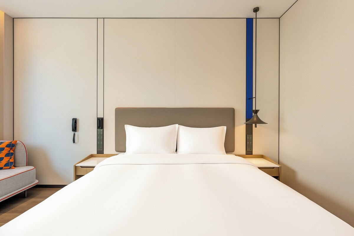 Ett hotellrum med en säng, ett sängbord, en lampa, en telefon och en soffa med en mönstrad kudde.