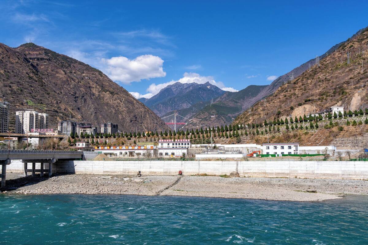 A river with a bridge, mountains, and buildings along the riverbank.