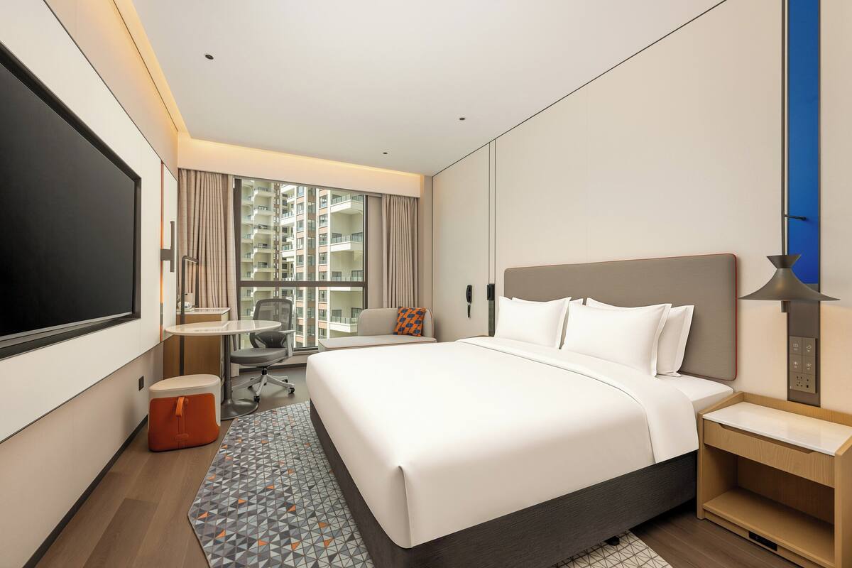 A hotel room with a large bed, a desk, a chair, a small table, and a view of the outside.