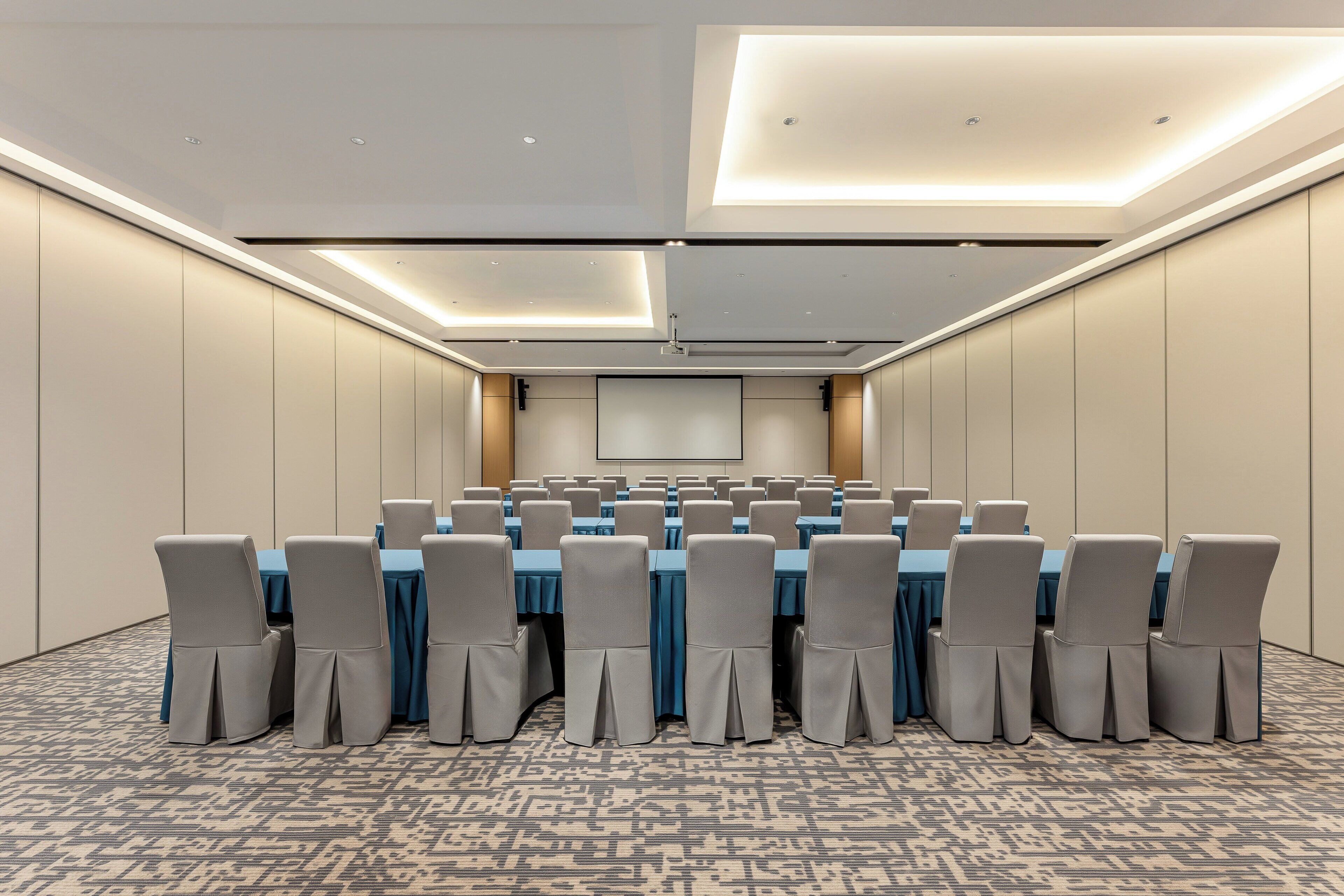 Un salón de conferencias con varias mesas y sillas, una pantalla de proyector e iluminación empotrada.