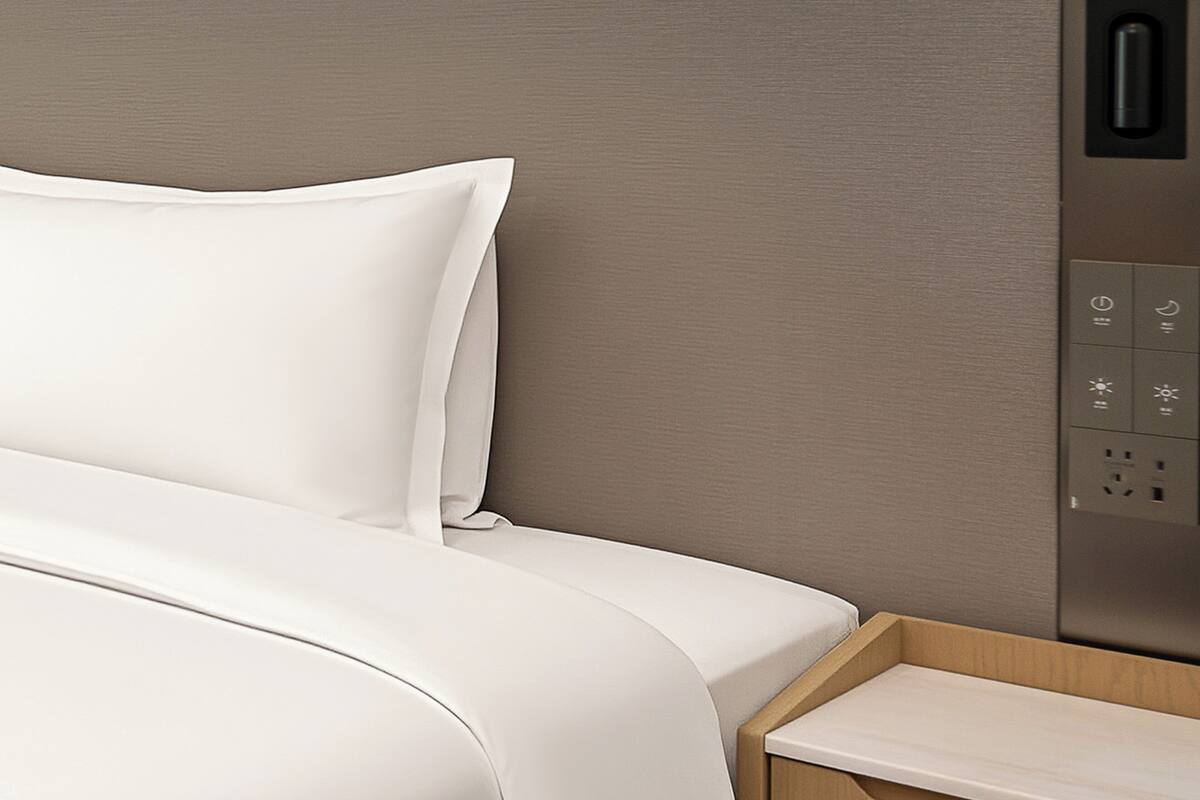 A bed with white bedding, a wooden nightstand, and a wall-mounted control panel with various buttons.