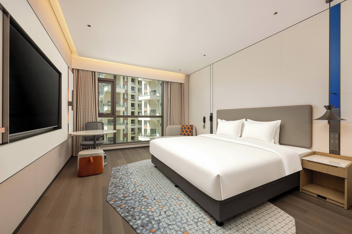 A modern hotel room with a large bed, a desk, a chair, a TV, and a view of the city.
