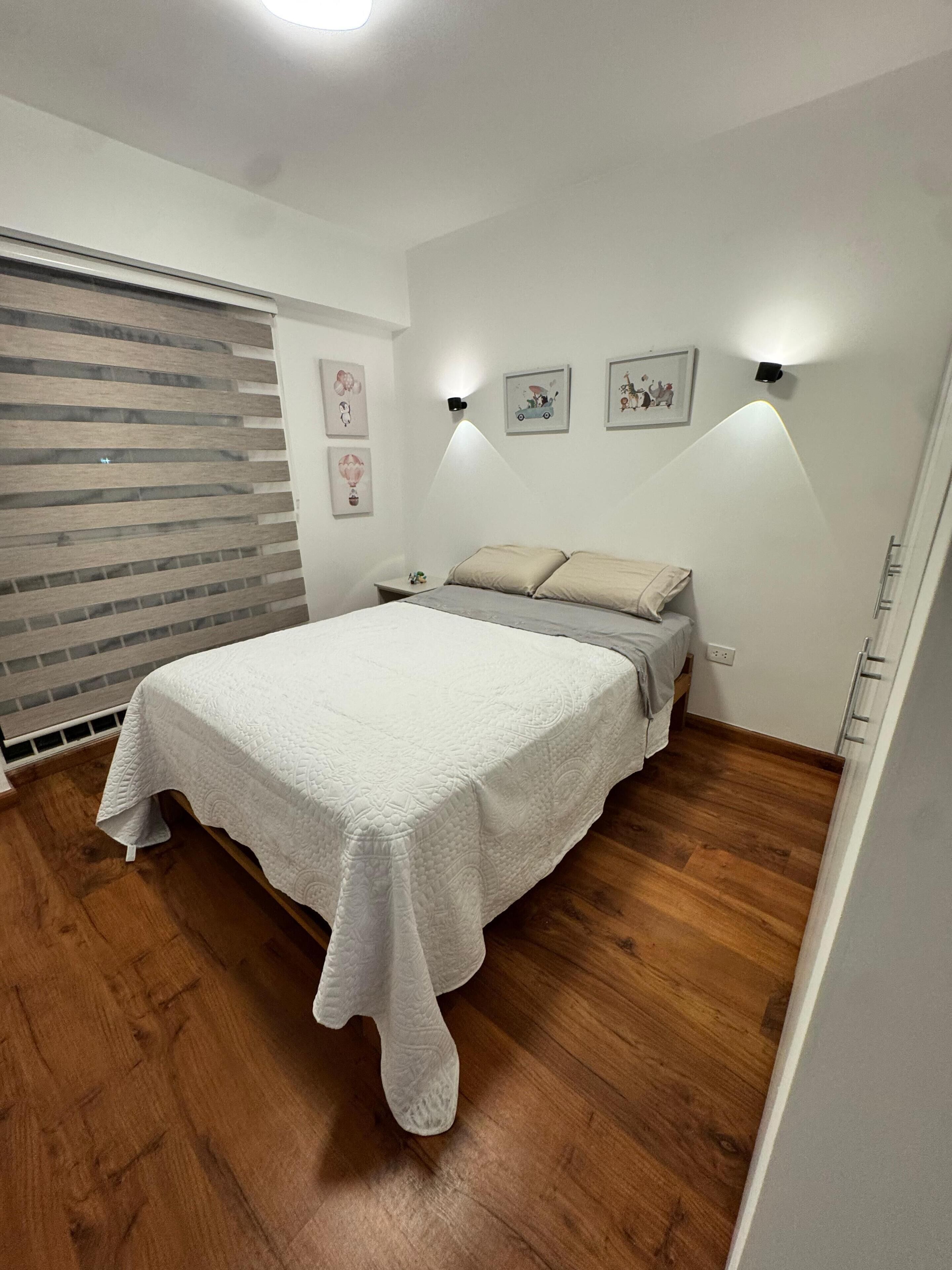 A bedroom with a bed, wooden flooring, a window with blinds, and wall-mounted lights.
