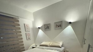 Quarto com cama, piso de madeira, mesa de cabeceira e iluminação na parede.