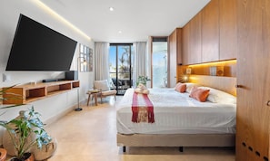 A modern bedroom with a large bed, a flat-screen TV, a balcony with a view, and wooden cabinetry.