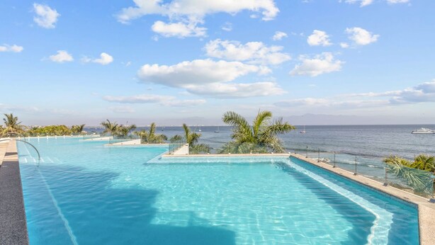 A pool with clear blue water, surrounded by palm trees and a view of the sea. - Zantamar 104B: Beachfront Studio Steps to Ocean (Cruz de Huanacaxtle)
