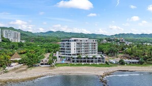 A modern hotel with multiple floors, a swimming pool, and a view of the surrounding mountains and beach. - Zantamar 104B: Beachfront Studio Steps to Ocean (Cruz de Huanacaxtle)