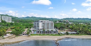 A beachfront hotel with multiple floors, a pool, and a view of the surrounding hills and greenery.