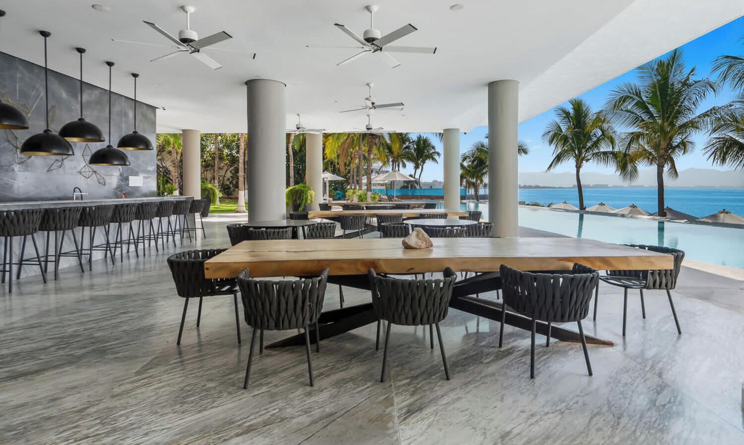 Un vaste espace extérieur aménagé pour les repas, avec une longue table en bois et des chaises noires, entouré de grandes colonnes et de ventilateurs de plafond.