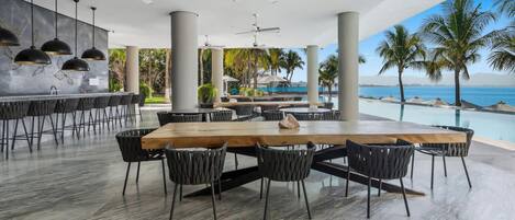 Un vaste espace extérieur aménagé pour les repas, avec une longue table en bois et des chaises noires, entouré de grandes colonnes et de ventilateurs de plafond.