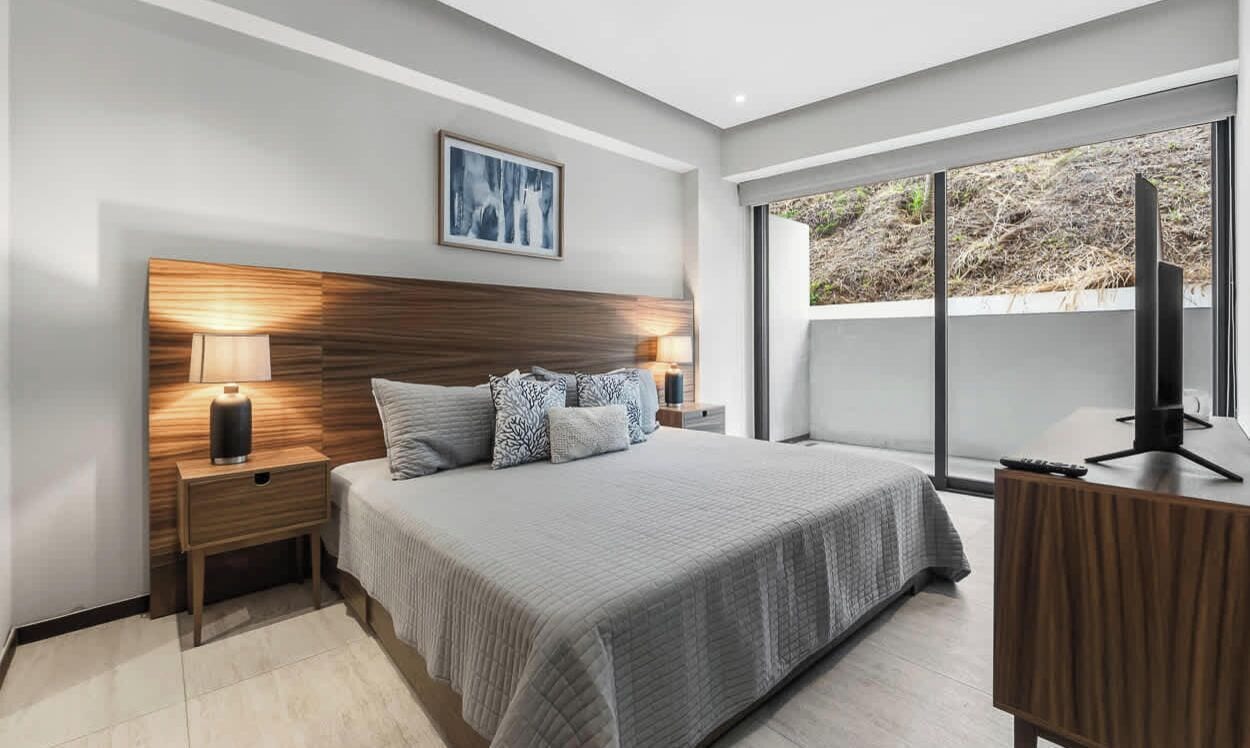 A modern bedroom with a large bed, bedside lamps, a wooden headboard, a desk with a chair, and a large window with a view.