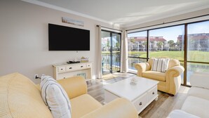 Un salon spacieux doté d’une grande porte-fenêtre coulissante donnant sur un balcon, d’un téléviseur à écran plat fixé au mur et d’une table basse blanche.