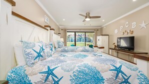 Une chambre à coucher avec un lit orné d’un motif représentant des étoiles de mer et des coraux, un ventilateur de plafond, une télévision et un balcon offrant une vue sur l’extérieur.