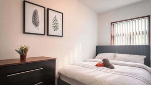 Une chambre à coucher comprenant un lit, une commode, une plante en pot et deux reproductions botaniques encadrées accrochées au mur.