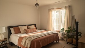 A bedroom with a bed, a bedside table, a lamp, a mirror, a window with curtains, and a plant. - Delightful 4-bedroom Plus House in  East Chatham with Pool, Sauna and AC (East Chatham)