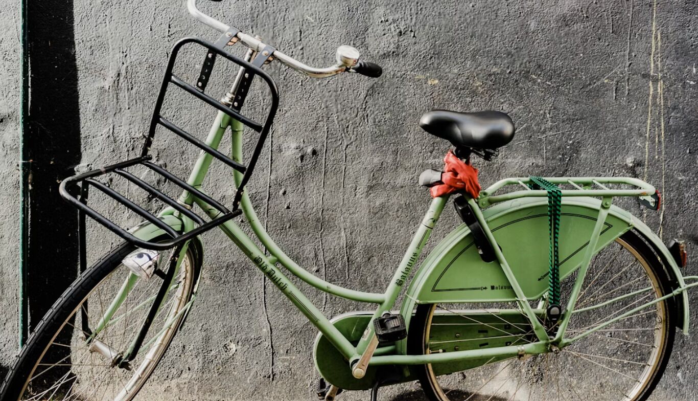 A green bicycle with a basket and a red bag attached to the rear, parked against a textured wall.