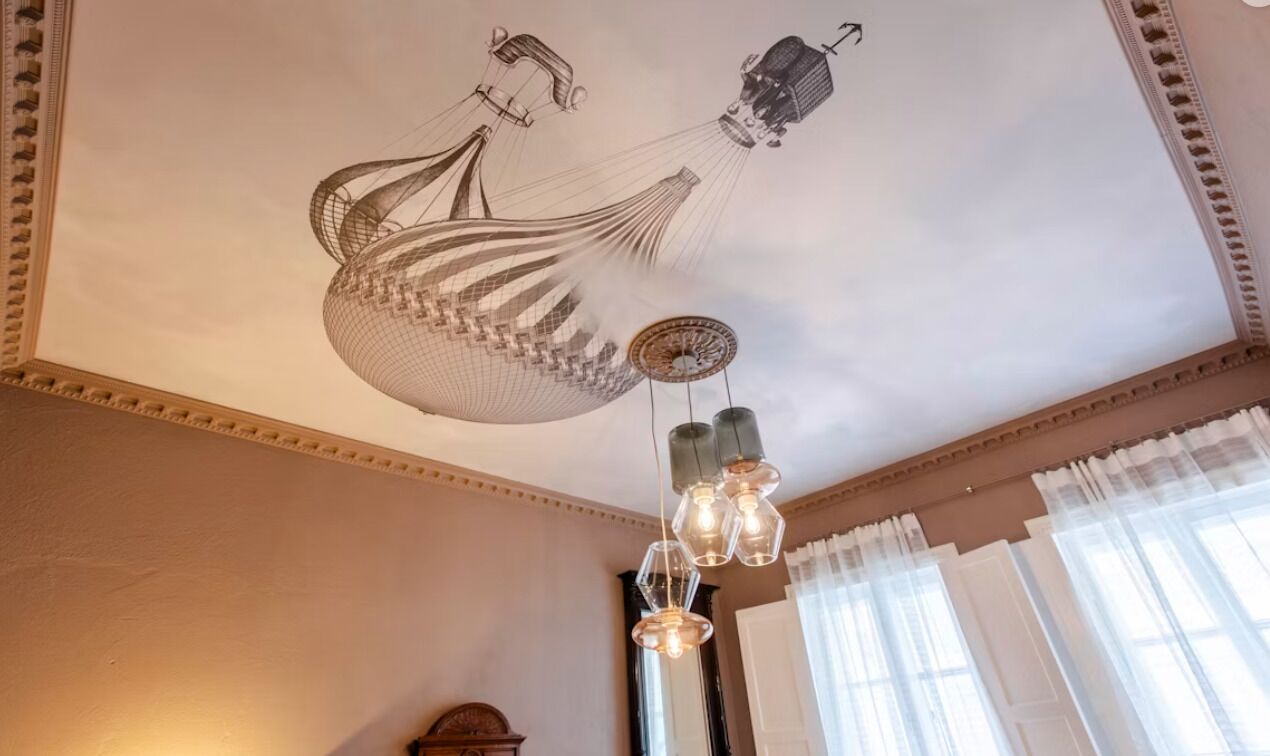 Une pièce avec un plafond haut, un lustre et une grande fenêtre avec des rideaux.