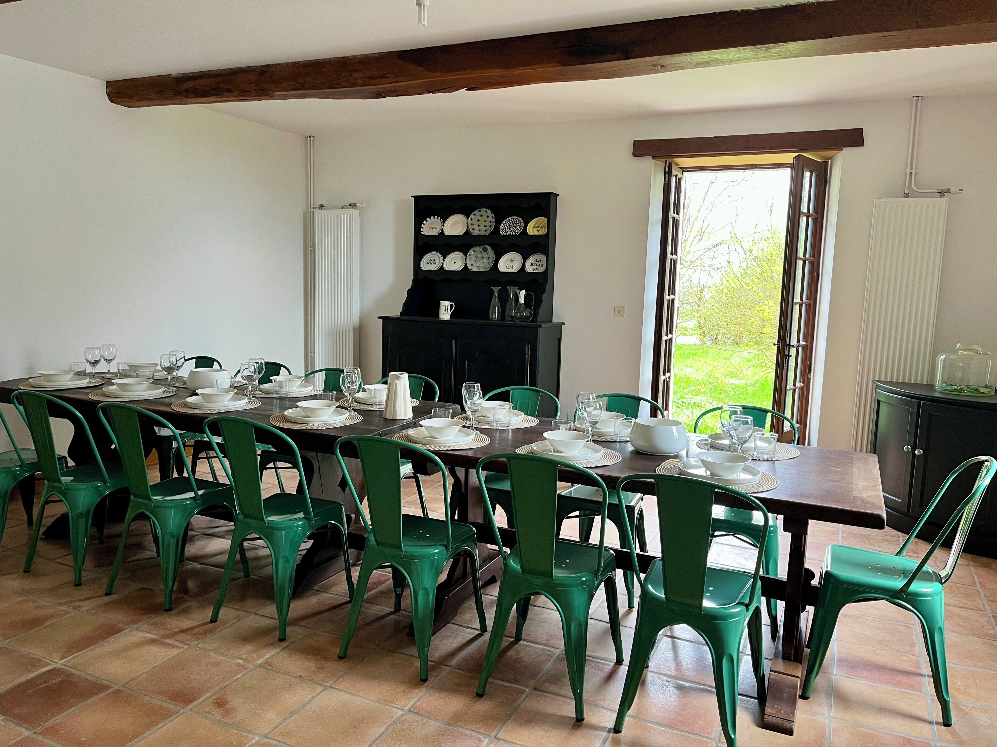 Un espace repas avec une longue table dressée pour un repas, entourée de chaises vertes, d’un buffet noir contenant de la vaisselle et d’une fenêtre donnant sur un paysage verdoyant.