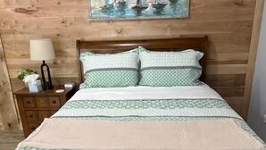 A wooden bed with a patterned bedspread and pillows, a nightstand with a lamp, and a painting of sailboats on the wall.