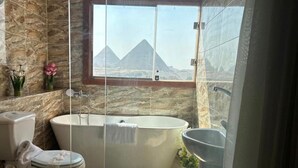 A bathroom with a bathtub, toilet, and sink, with a view of pyramids through a window. - Spacious 9-bedroom hotel in charming Giza Governorate with AC (Giza Governorate)