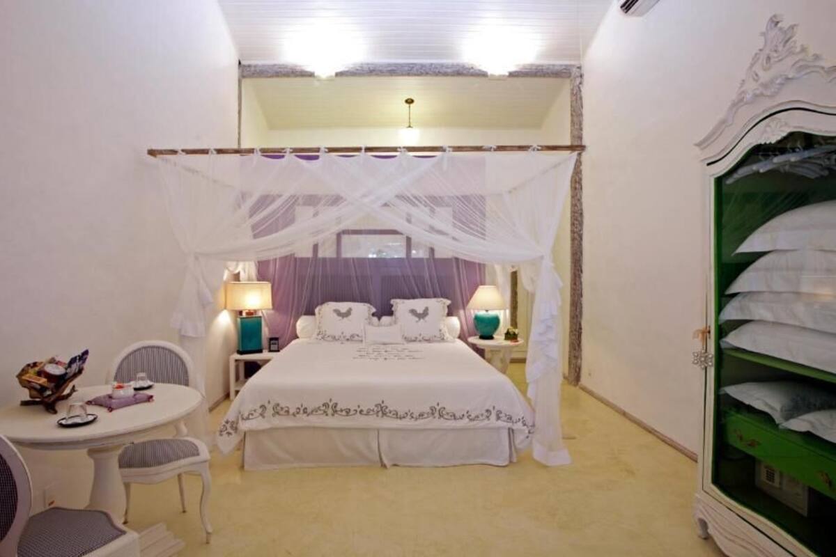 A bedroom with a canopy bed, bedside lamps, a small round table with chairs, and a wardrobe with green shelves.