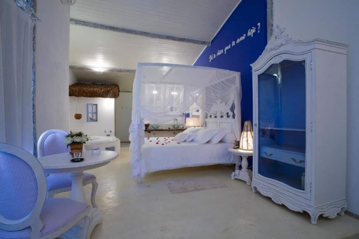 A bedroom with a four-poster bed, a bedside table with a lamp, a white cabinet, and a chair.