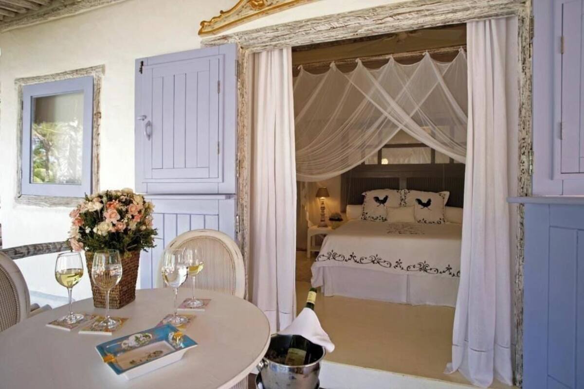 A room with a bed, a table with wine glasses, a basket of flowers, and a window with curtains.