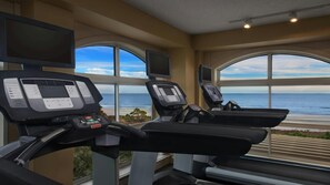 Un gimnasio con cintas de correr y equipo de ejercicio, ventanas amplias con vista al océano e iluminación empotrada en el techo.