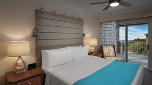 Een hotelkamer met een bed, twee bedlampen, een nachtkastje, een stoel, een balkon met uitzicht en een plafondventilator.