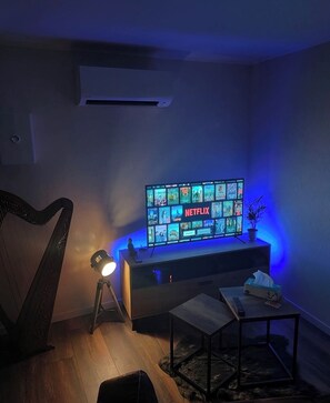 Ein Wohnzimmer mit Fernseher, der eine Netflix-Oberfläche anzeigt, einer Lampe, einem kleinen Tisch und einer Harfe.