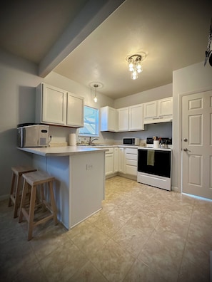A kitchen with white cabinets, a microwave, a stove, and a sink. There are wooden stools and a ceiling light fixture. - Cozy & Convenient Casper Stay (Casper)