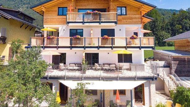 A two-story wooden building with balconies, outdoor seating, and a mountain backdrop. - Apartment Multicolor Amethyst at Wolfgangsee Alpenblick (St. Wolfgang)