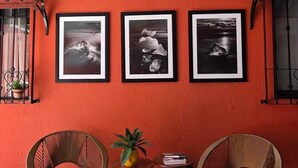Una habitación con una pared roja, tres fotografías enmarcadas, dos sillas de mimbre, una mesita con una planta y libros, y una ventana con vistas.