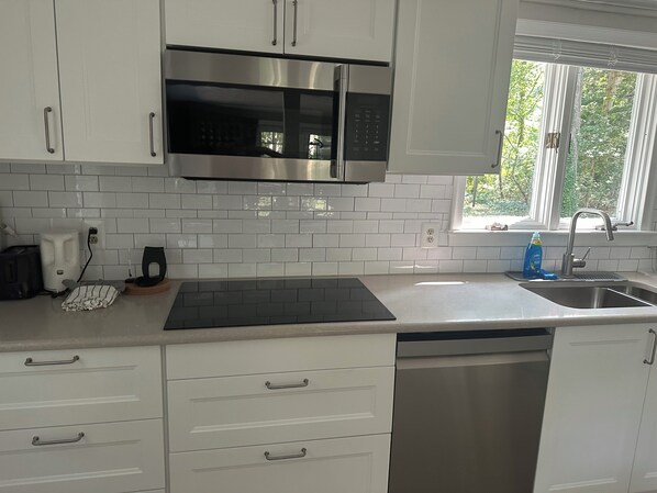 Cucina con mobili bianchi, un forno a microonde in acciaio inossidabile, un piano cottura a induzione nero, una lavastoviglie e una finestra vista verde.