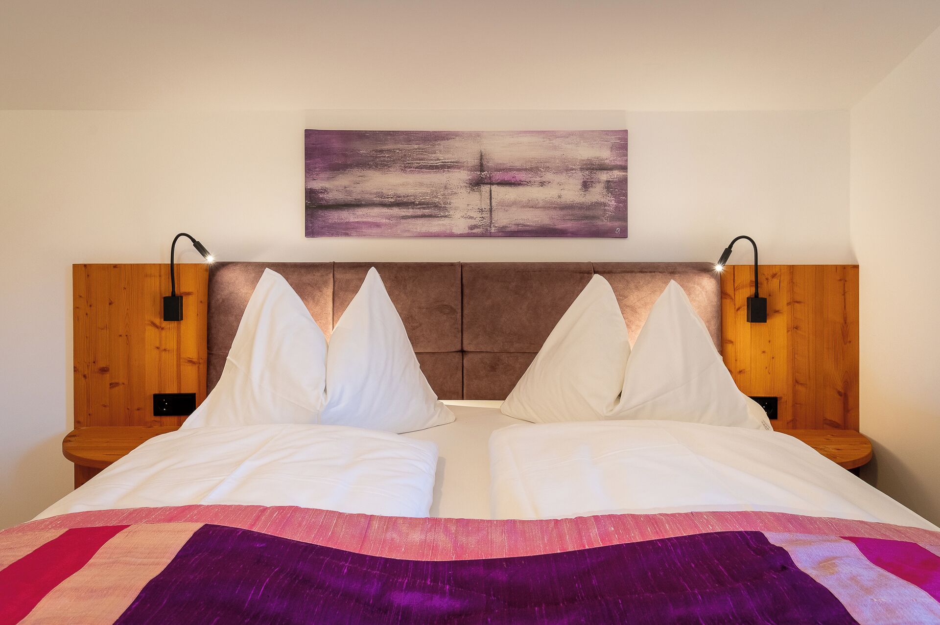 A bed with a headboard, two bedside tables, a wall-mounted light, and a painting above the bed.