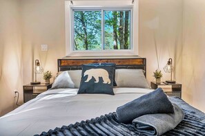 Ein Schlafzimmer mit einem großen Bett, einem Fenster mit Blick auf Bäume, Nachttischen mit Lampen und einem Kissen mit Bärenmotiv.