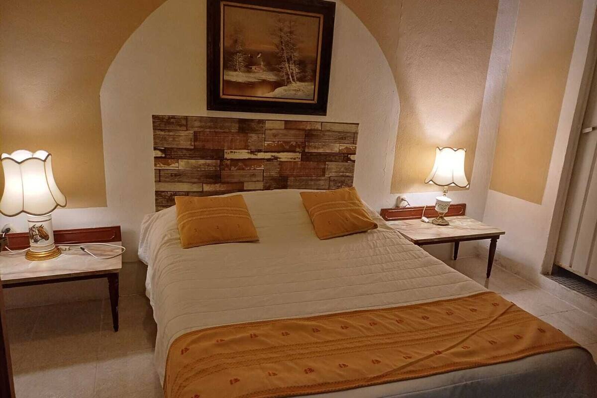 Un dormitorio con cabecera de madera, dos mesitas de noche con lámparas, una cama con colcha blanca y dos almohadas, y un cuadro sobre la cabecera.
