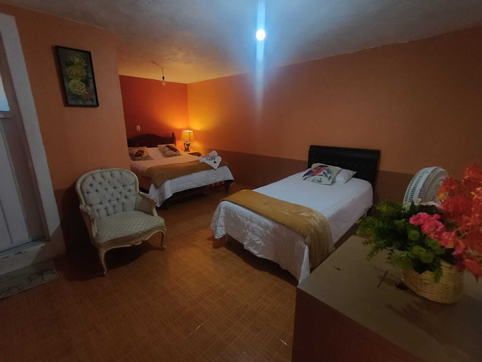 A hotel room with two beds, a chair, a bedside table, a lamp, and a fan.