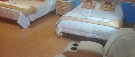 Habitación de hotel con dos camas, una silla de masaje, una mesita de noche con lámpara y decoración floral en la pared.