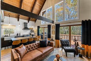 Een ruime woonkamer met een grote bruine leren bank, een eettafel met stoelen, een keuken met een bar en grote ramen met uitzicht op bomen.